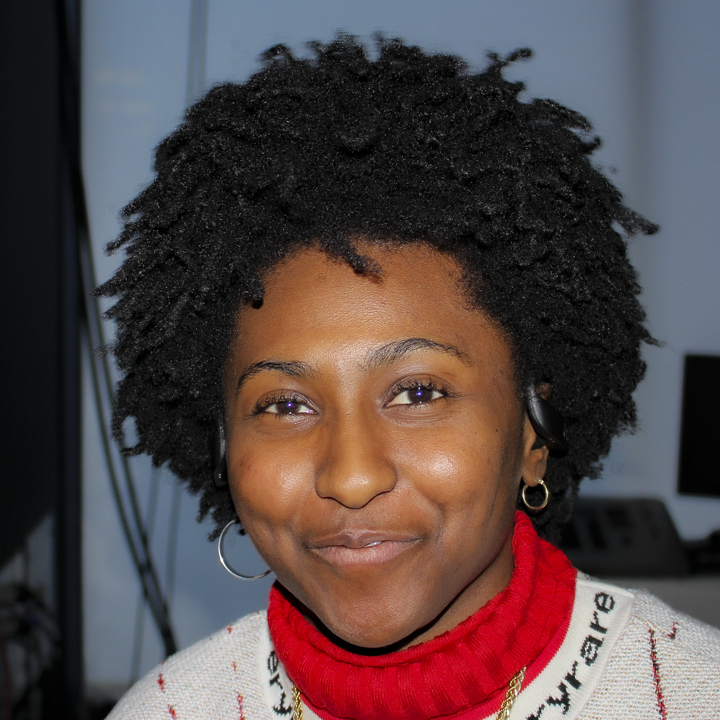 A barefaced melanated person smiling with an afro, asymmetrical hoops, gold chains, red turtleneck, and a white jacquard knit.