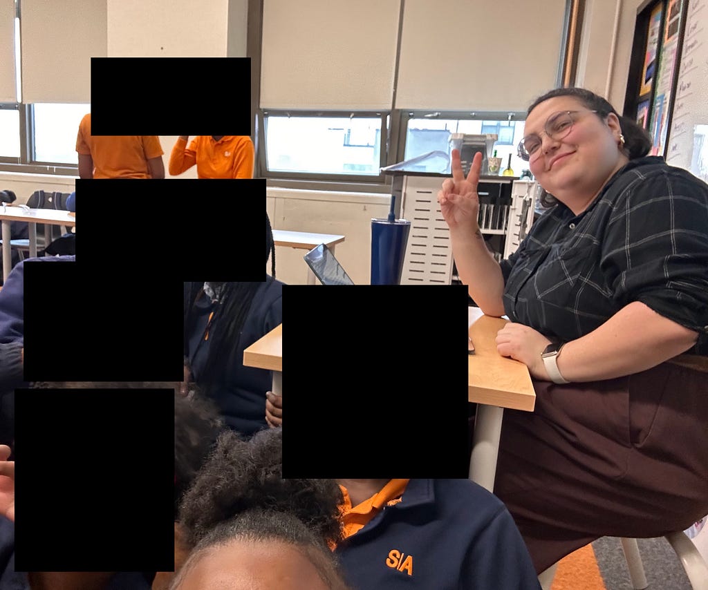 Me, the author, sitting at a desk, smiling and holding up a peace sign. Students, also in the picture, have their faces censored, but the SA logo is visible on their shirts and sweaters.