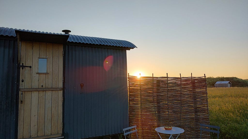 A partial view of a shepards hut made of metal frame and light wood door. There is a privacy screen / fence made of rattan or bambo like material and behind it you can see green grass and a setting sun