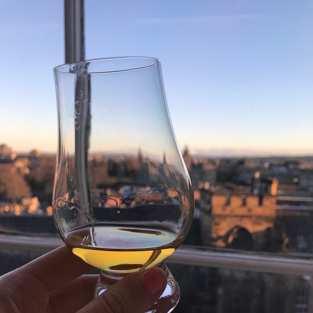 Whisky glass with a small amount of whisky held up next to a large window and the city of Edinburgh, Scotland as the blurred background. The city is enveloped in a warm, golden hue from the light of the setting sun.