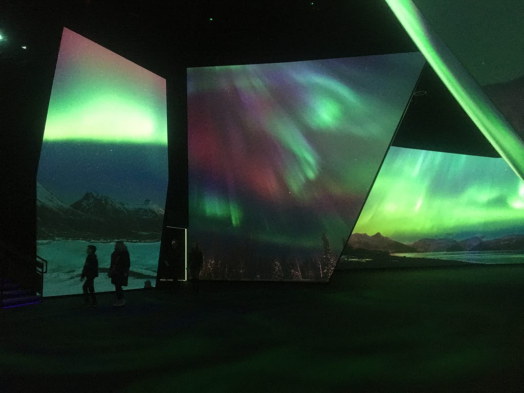 In a large, dark room, a series of enormous screens of different shapes feature a live-action footage of the aurora borealis from the BBC’s Antartica episode of Seven Worlds, One Planet