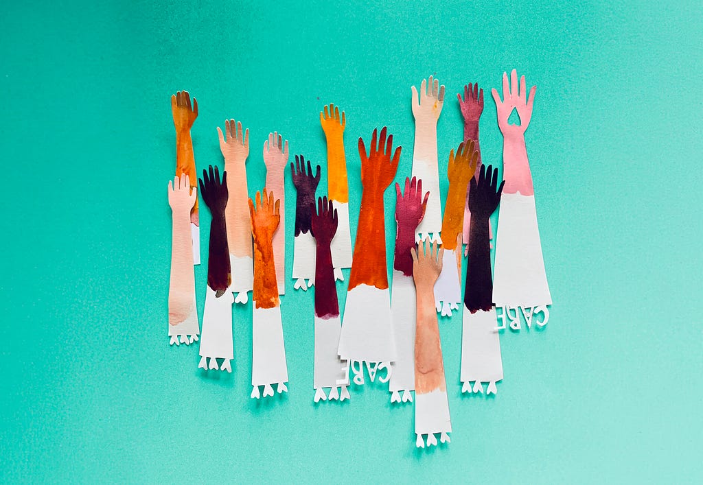 Colorful paper cutouts of raised arms in various skin tones, symbolizing diversity and inclusion, against a teal background. The bottom of each paper arm features cutouts of hearts and the word “CARE.”