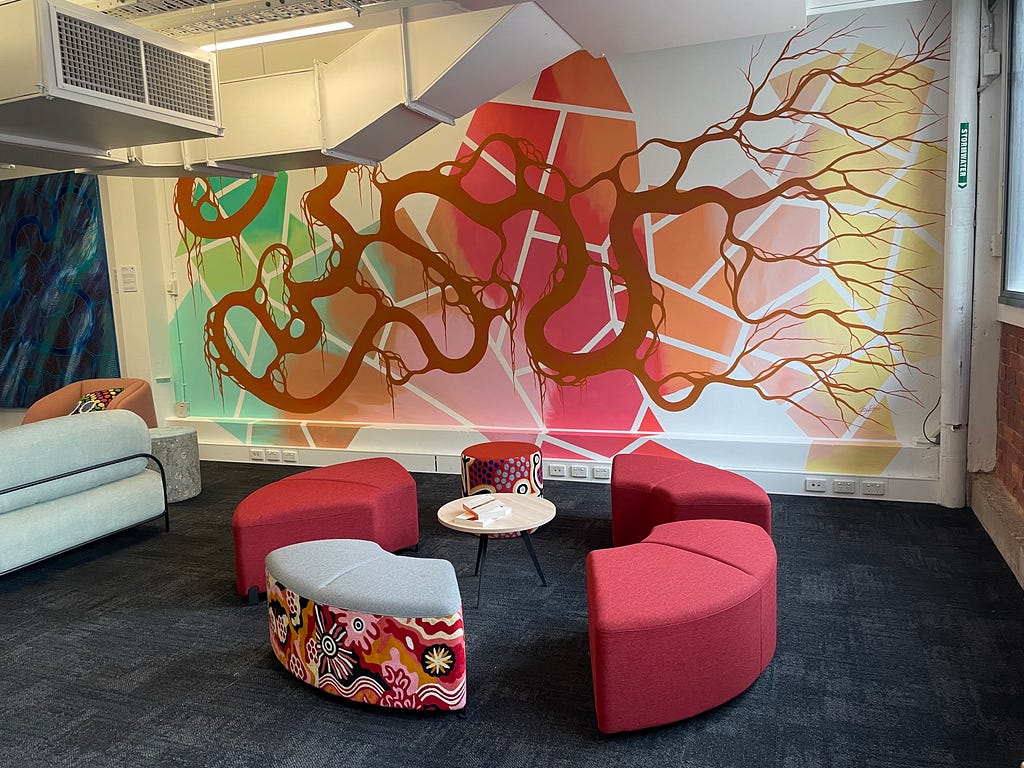 Bright coloured mural with geometric shapes in the background and organic twisting form in the foreground by Troy Firebrace. Furniture with Indigenous designs by WINYA located in front of the mural.