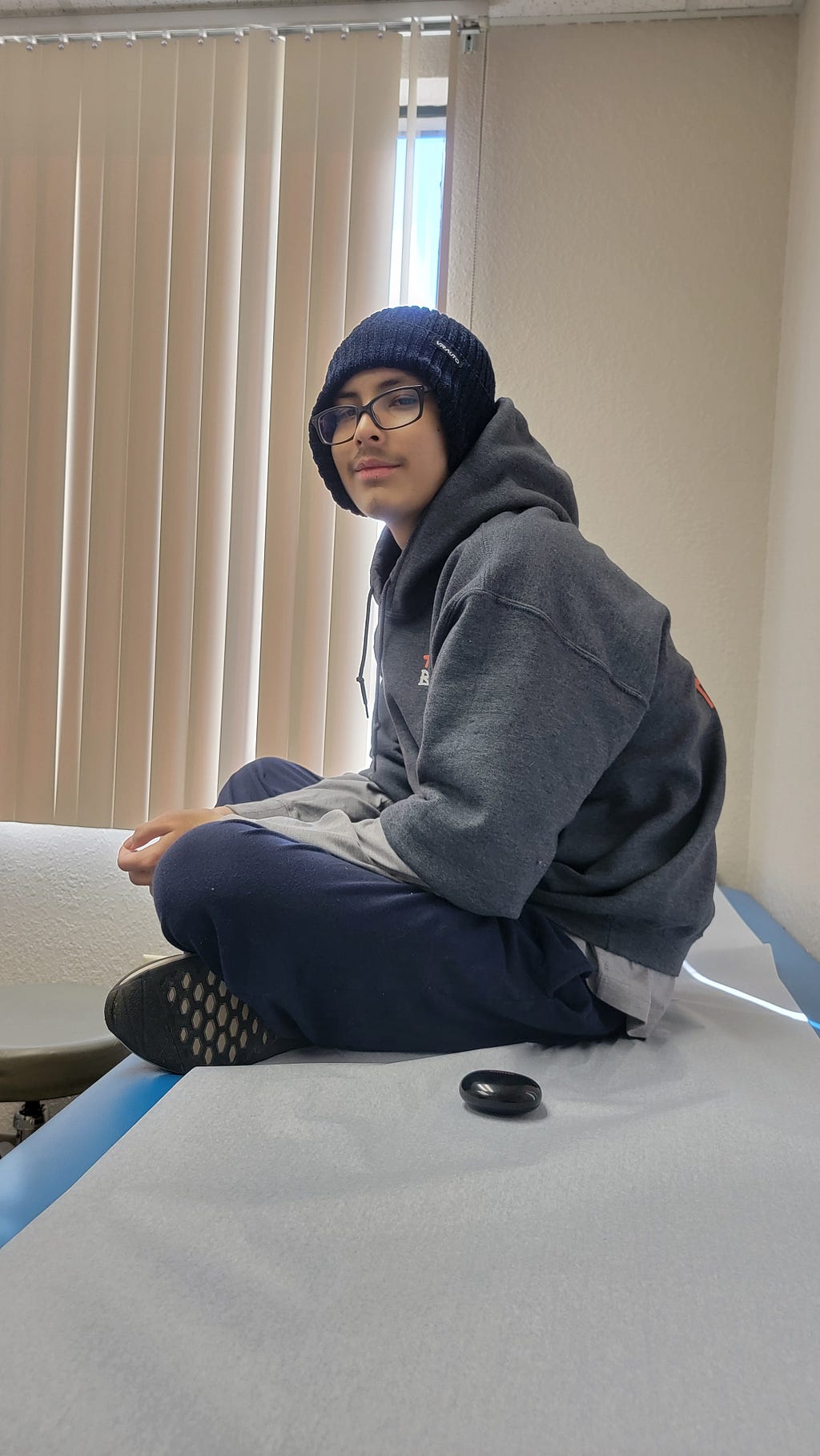 A person wearing glasses, a beanie, and a cozy gray hoodie sits cross-legged on a medical examination table, conveying a calm and relaxed mood.