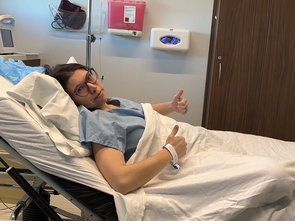 Bri in a blue hospital gown, laying in a hospital bed and covered with white blankets. She is giving two thumbs up and a smile before her egg retrieval procedure.