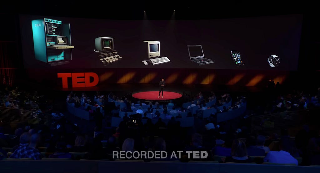 Imran Chaudhri presenting the Humane AI pin concept on the TED stage with the background slide showing an image of evoluation of computers from large size PCs, to laptops, to phones to a smartwatch now.