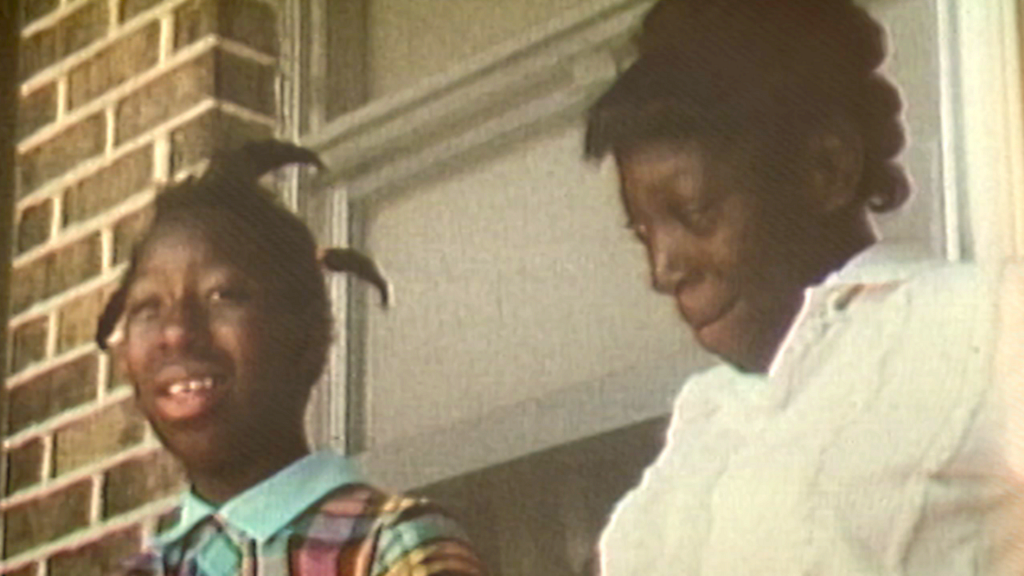 Mary Alice and Minnie Lee Relf sit on a porch, smiling. One is dressed in white, the other in a colorful blue shirt.