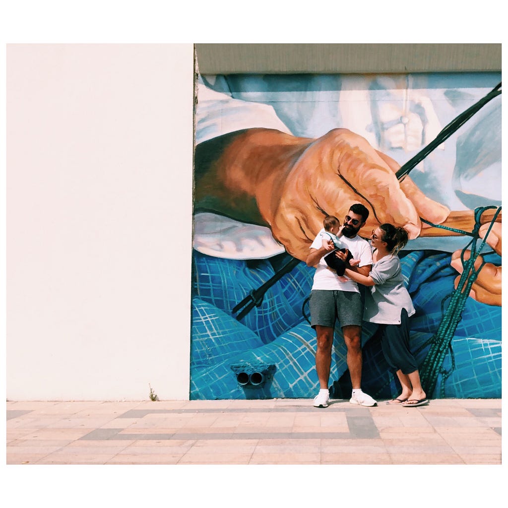 My husband carries our son and I’m leaning over and hugging them from the side. We are standing in front of a wall mural.