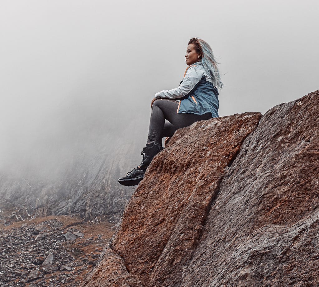person with long light blue dyed hair and wearing a sporty jacket and boots sitting on a rock ledge in profile with more rocky landscape and fog behind