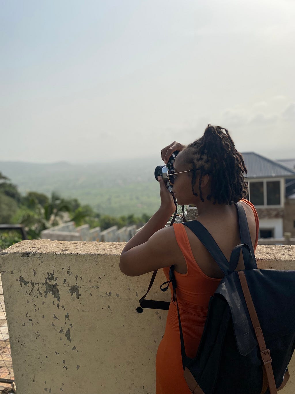 Woman with short locs, sunglasses and a backpack stands sideways, looking into a camera. She is taking a photo of the green Aburi mountains in Ghana.