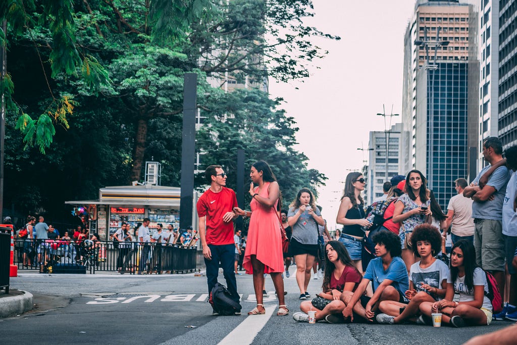 Muitas pessoas na Avenida Paulista, próximo ao Parque Trianon; algumas em pé conversando e outras sentadas.