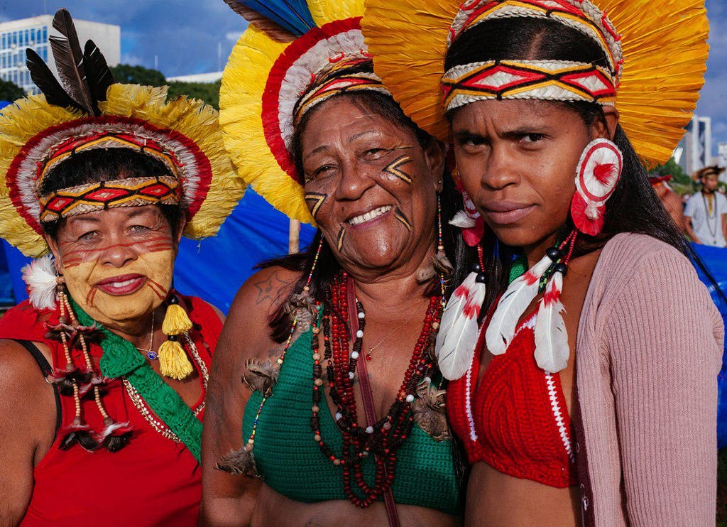 Três mulheres indígenas olha para a lente da câmera. Duas são mais velhas e sorriem. A mais jovem parece desconfiada