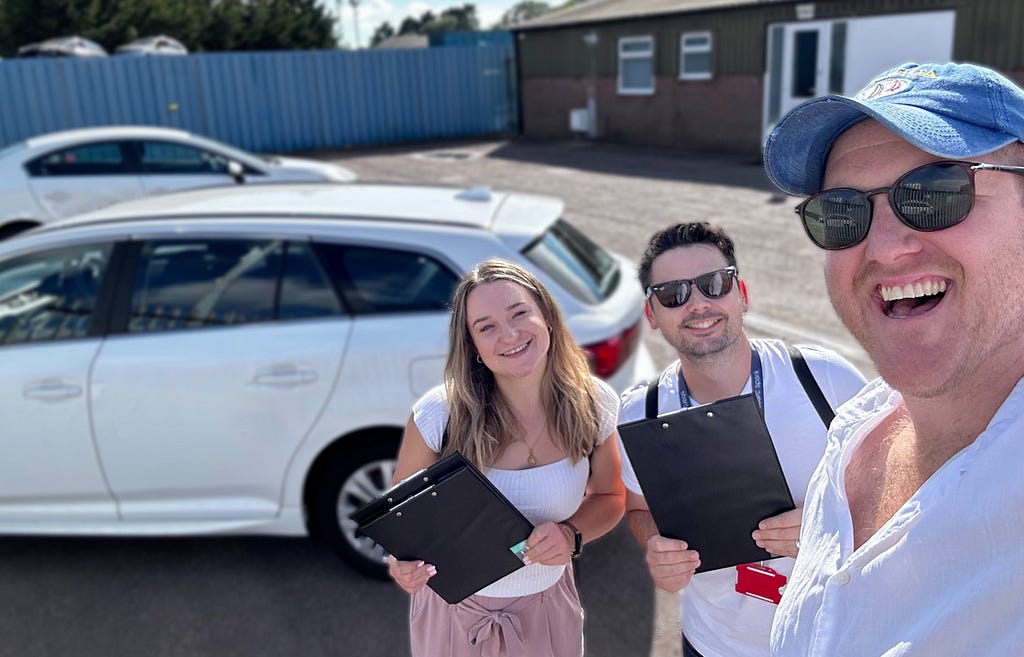 Three people standing by a parked car, smiling at the camera. They are holding clipboards and appear to be conducting surveys or interviews. A white car is visible in the background, along with some buildings. The image has a casual, outdoor work setting feel to it.