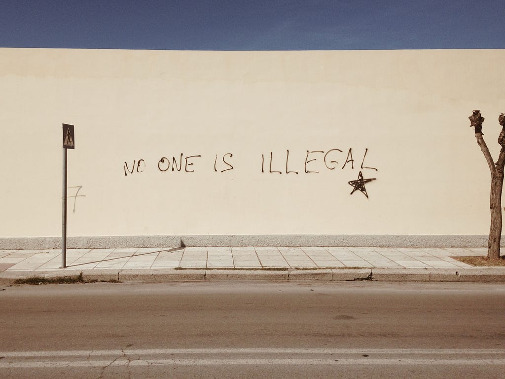 A white wall with the phrase “no one is illegal” written in spray paint