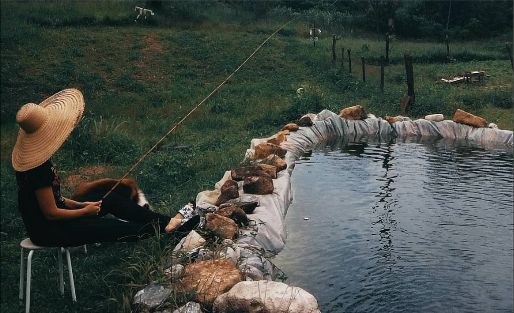 Imagem que contém um tanque de peixe e uma pessoa sentada ao lado enquanto pesca, em um sítio.