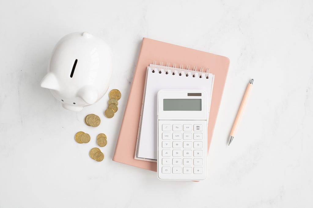 marble desk flatlay with white piggy bank, peach notebook, gold coins and calculator