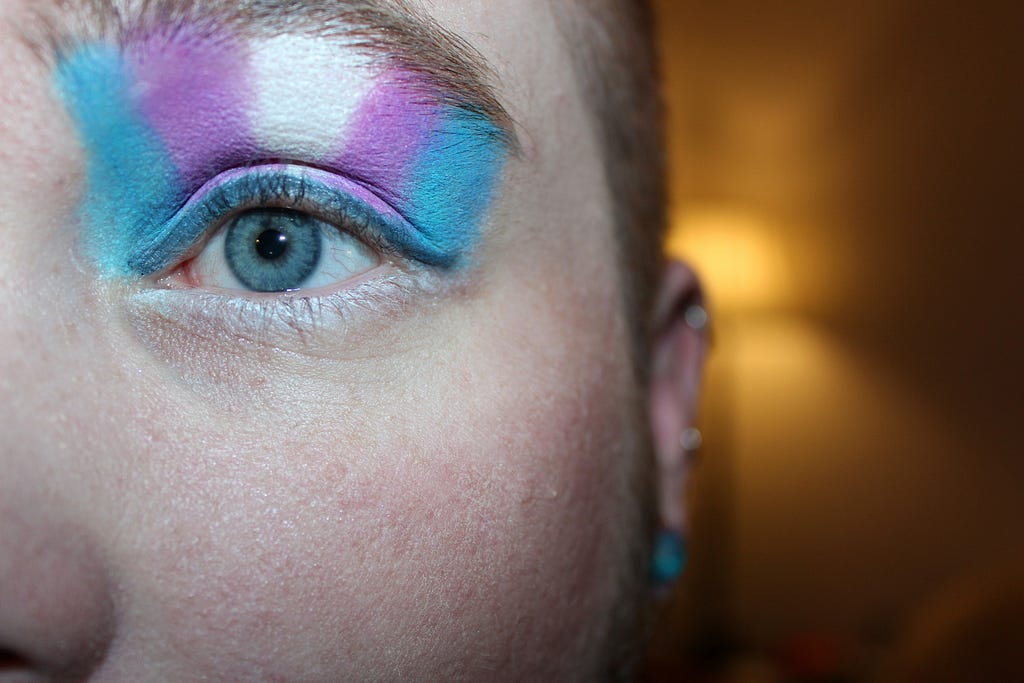 Close up photograph of half of a face of a caucasian youth, wearing studs earrings, and shorn dirty blond hair. The photo is cenbtered on their half of their face, one blue eye and eyebrow. Above the eyebrow is the trans flag in eyeshadow extending to the lower eyebrow.