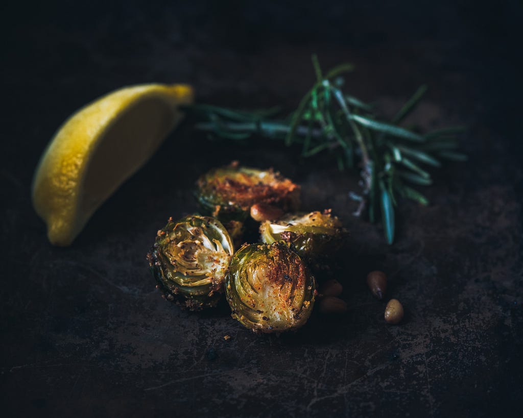 Roasted brussels sprouts on a dark stone background. Sliced lemon and rosemary twigs lay beside it.
