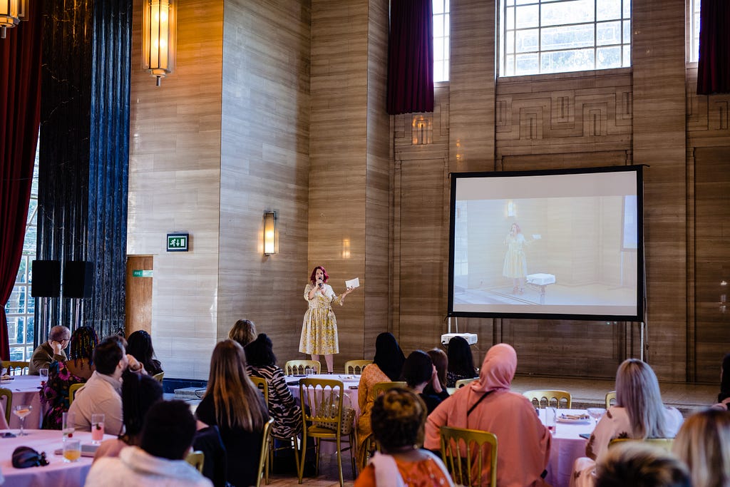 A white woman in a floral dress speaks into a microphone on a stage, with the audience in front of her and a screen to her right showing a live feed of the event.