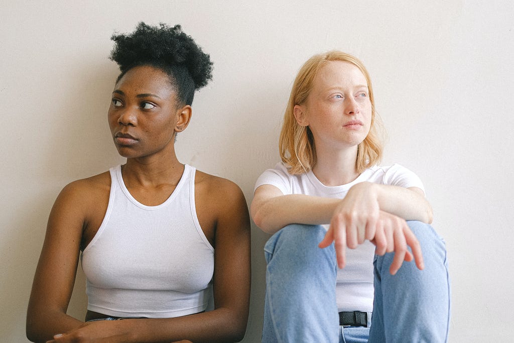 Two women seated, one White and one Black, looking vaguely unhappy.