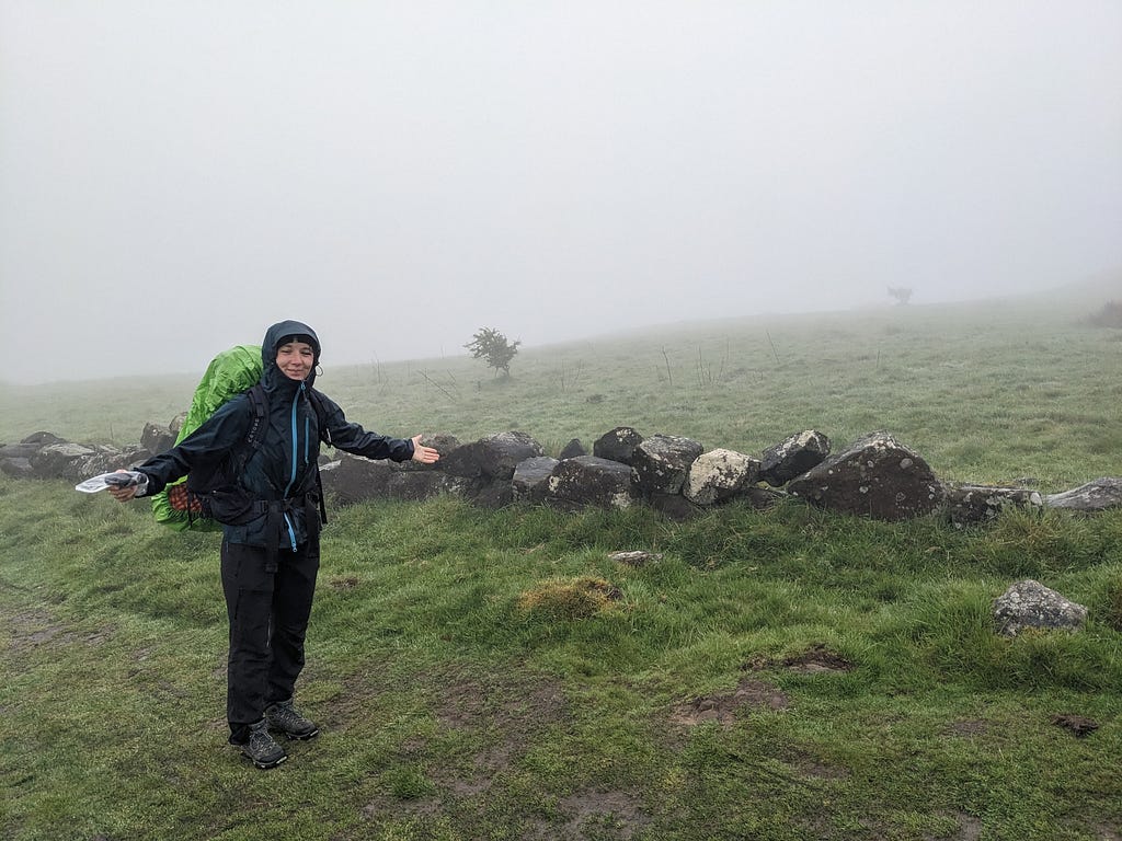 Caitlin stands to the left of the picture in a desolate looking moorland that is shrouded in mist. She is wearing a long waterproof coat with the hood up and holds her arms wide to show off the glorious landscape comically