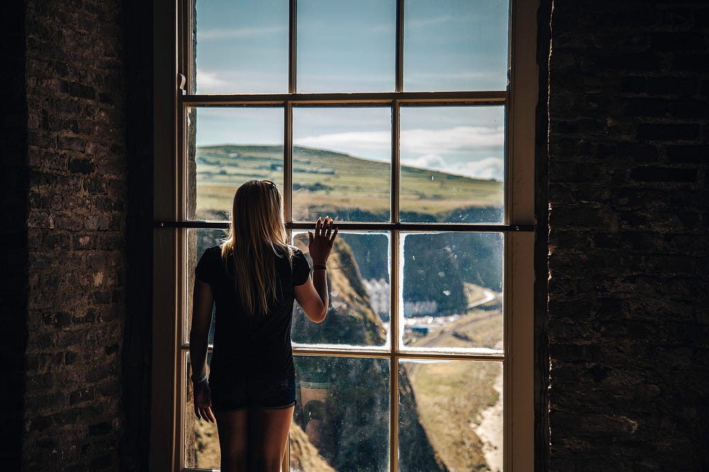 Woman looking through a pane glass window at a rural coastal landscape.