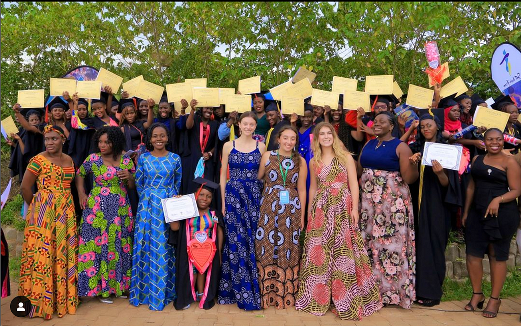 A large group of people standing together, wearing floor length dresses in multi-color patterns. The people in the back are all wearing caps and gowns and holding up certificates. Most everyone is smiling and looking at the camera.