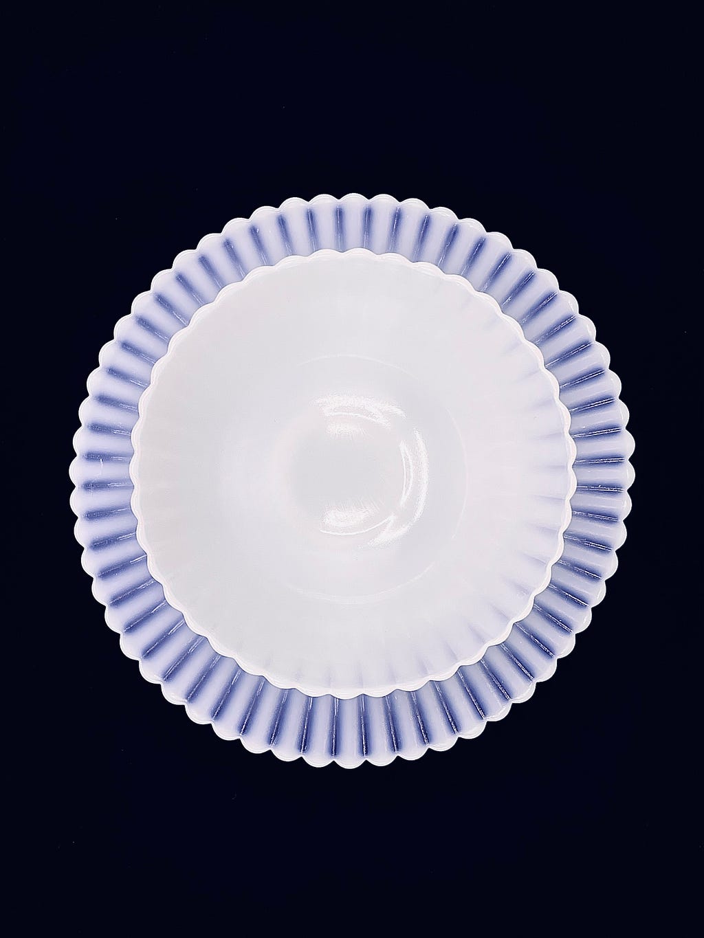 A top-down view of the compote dish stacked on top of the matching dessert plate against a black backdrop. The dish on top looks fully opaque while the visible margin of the plate beneath it glows purple at its thinnest parts.