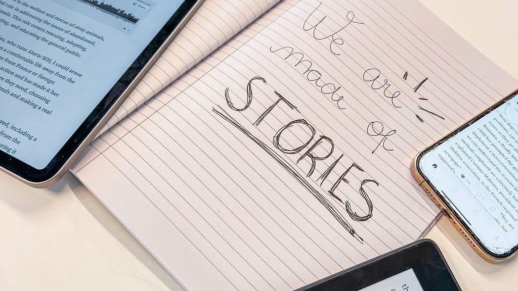 A photo of a notebook with the frase ‘we are made of stories’ written and an ipad, iphone and kindle showing articles.