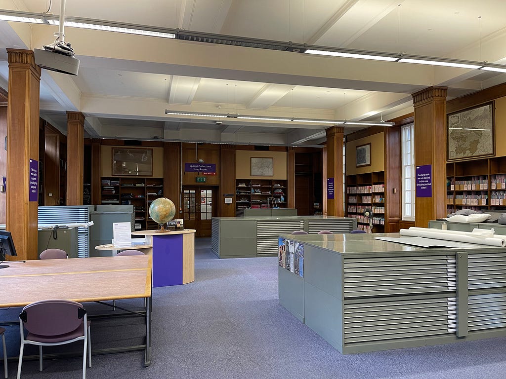 A photo of the Main Library’s Map room. In it, a table, a globe, two sets of large filing cabinets, and many shelves.