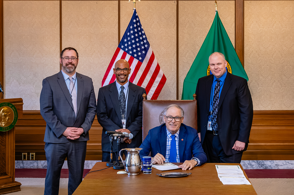 An image of LCB Legislative Relations Director Marc Webster, LCB Director Will Lukela, Gov. Jay Inslee (seated), and LCN Policy and External Affairs Director Justin Nordhorn in front of an American flag and a Washington State flag at the signing of HB 2260 in March 2024