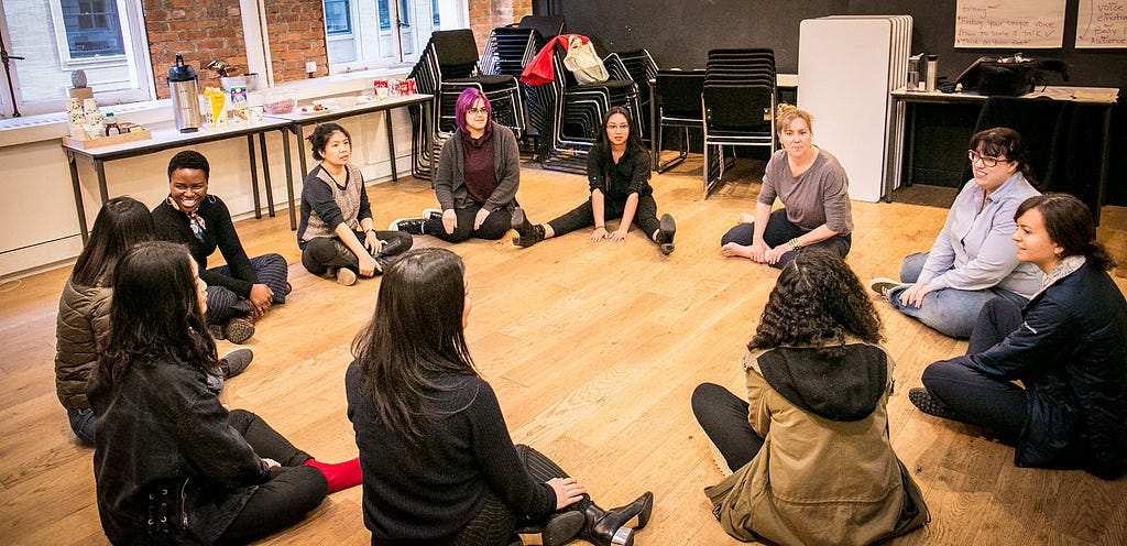 A group sitting on the wood floor in a classroom in a circle during a Women Talk Design speaking workshop