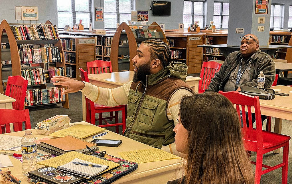 During the SJIEP 2024 event, a man with cornrowed hair and a vest gestures animatedly while speaking, capturing the attention of his colleagues seated at a library table with red chairs. Notebooks, pens, and personal items are strewn about, indicating an active discussion. In the background, another participant looks on attentively, embodying the collaborative spirit of the event.