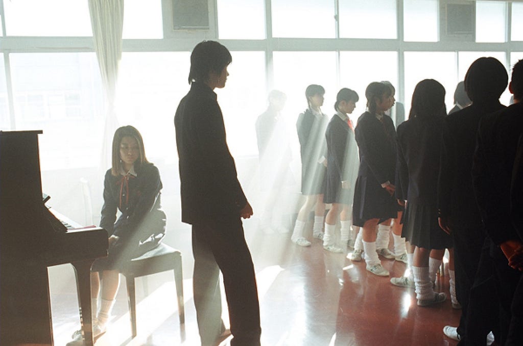 A choir room full of young teen boys and girls dress in black shirts and pants/skirts with white socks and shoes. To the right of the shot the choir is facing their back towards the composer and pianist to the left of the shot stands the composer and closely behind him the pianist who sits at a black grand piano. The choir room has an auburn marble floor and a wall of windows to the back that pours in soft white light from above which almost engulfs the room.