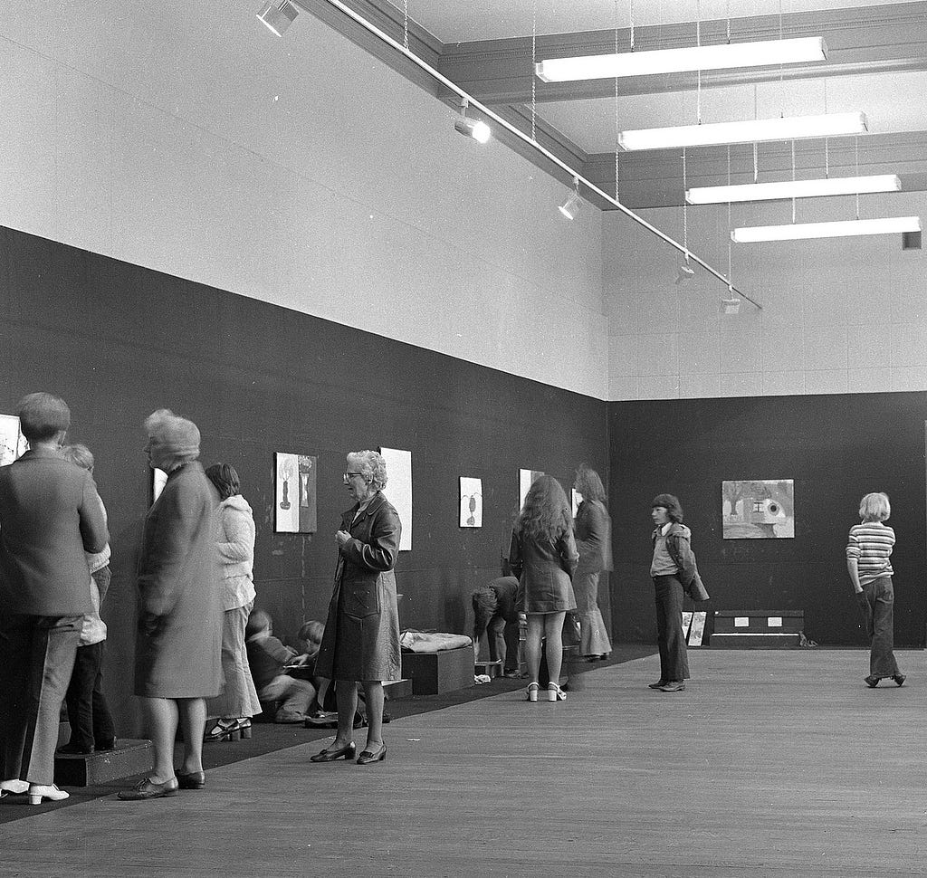 A black and white photo showing a scattered crowd looking at some artworks hung on a gallery wall