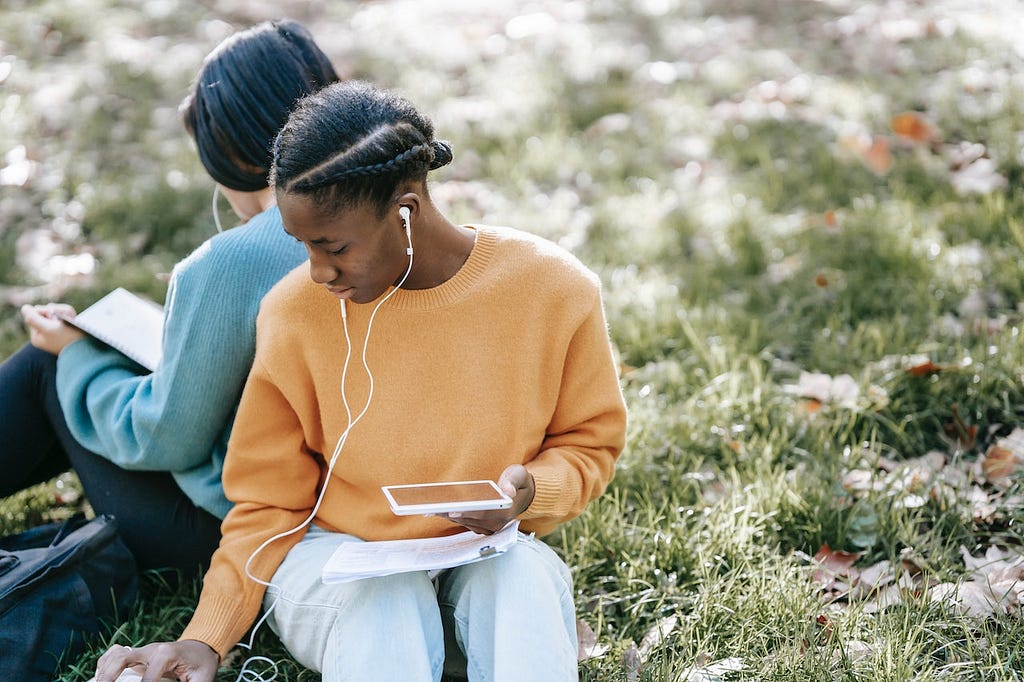 Two ladies seating on grass listening to their smartphones