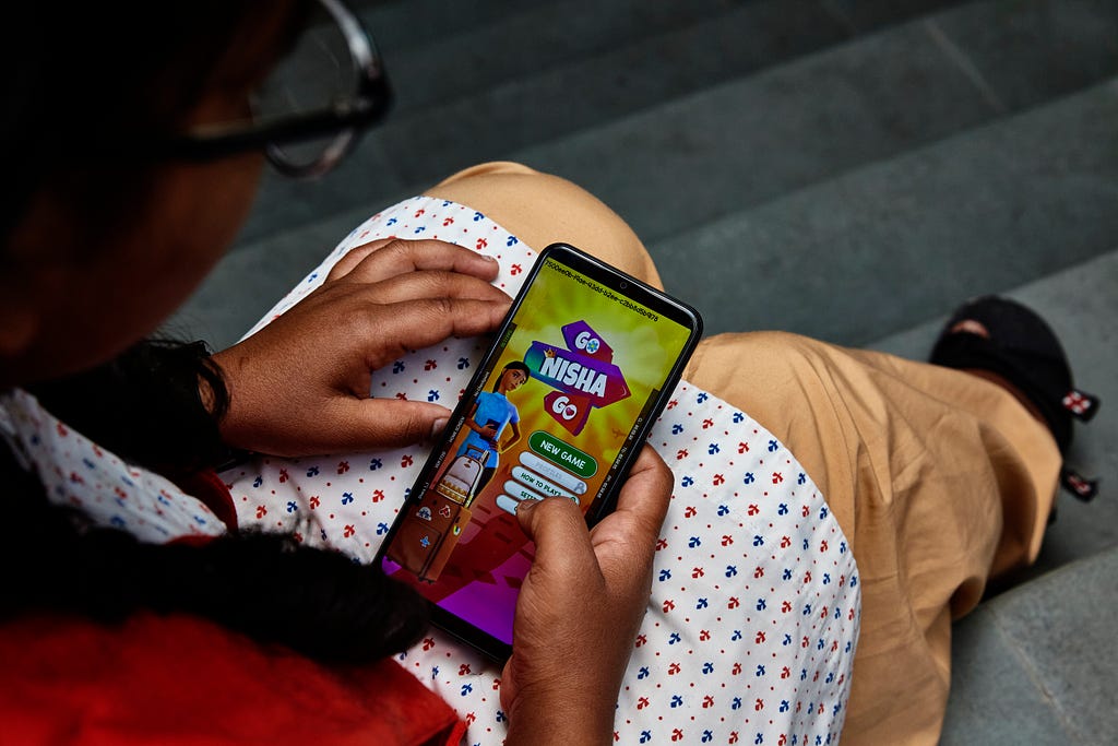 A girl holds a mobile phone in her lap with the game Go Nisha Go on screen and her thumb hovering over one of the buttons.