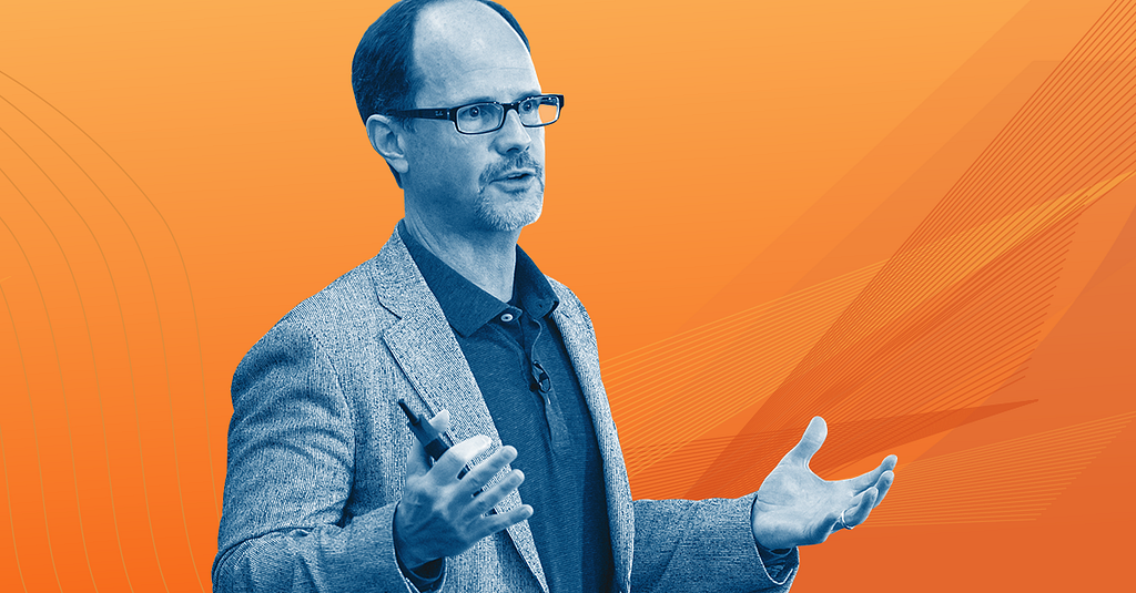 blue-scale photo of Rob Alexander wearing a blazer and gesturing mid sentence against an orange background.