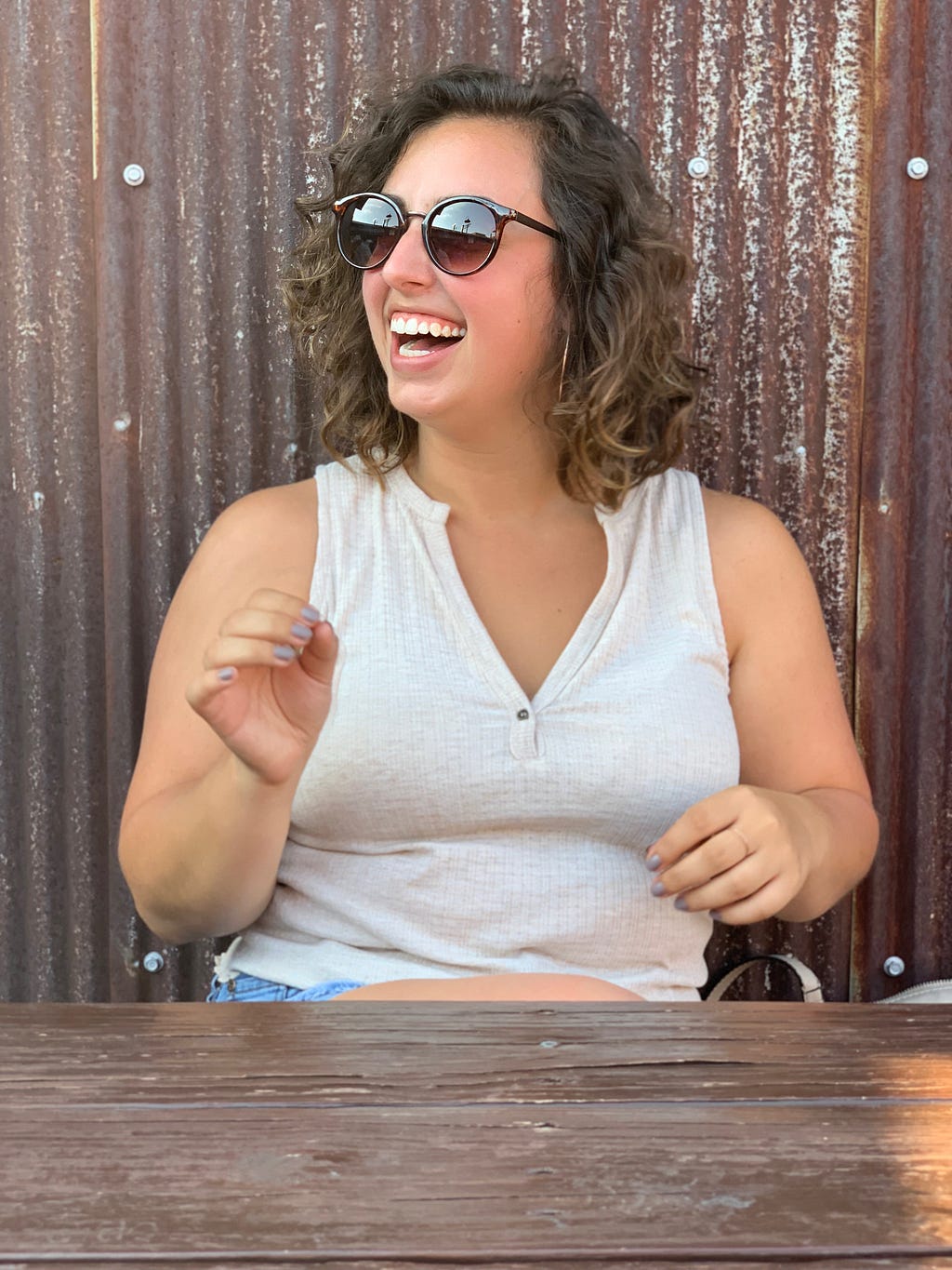 Young woman with short brown curly hair wears sunglasses and a big smile as she looks off to the side in front of metal.