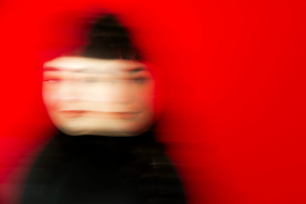 A blurred, artistic portrait of a person with dark hair against a vivid red background. The motion blur creates a dynamic, ghostly effect, with the face appearing multiple times in a horizontal direction.