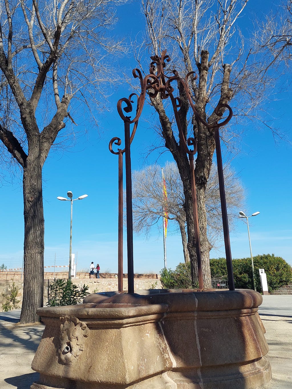 This image shows, in the foreground, a well surrounded by trees and benches, in the castle courtyard. In the background, you can see the flag of Catalonia, placed there by the Generalitat in 2011.