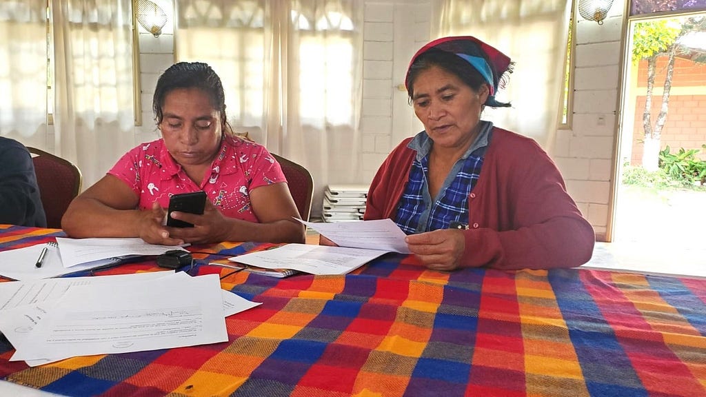Two women sit at a table with a colorful blue, red and orange checked table cloth while gazing down at a mobile phone and stacks of papers.