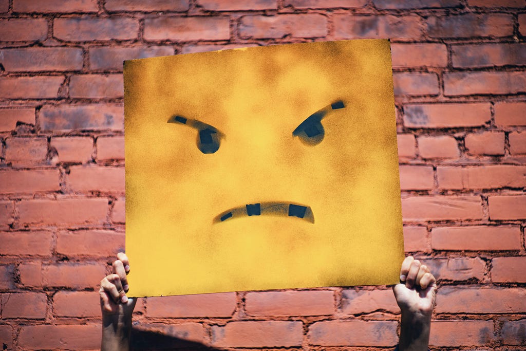 Hands holding a sign with a yellow angry face against a red brick backdrop