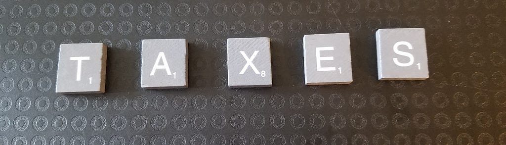 Grey wooden letter tiles on a black background. Letters spell TAXES.