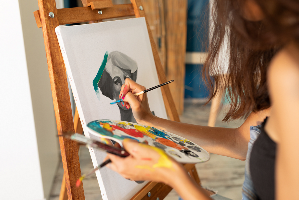 At the center of the photo is a painted portrait. A teenage girl’s hands can be seen holding paints and a paintbrush.