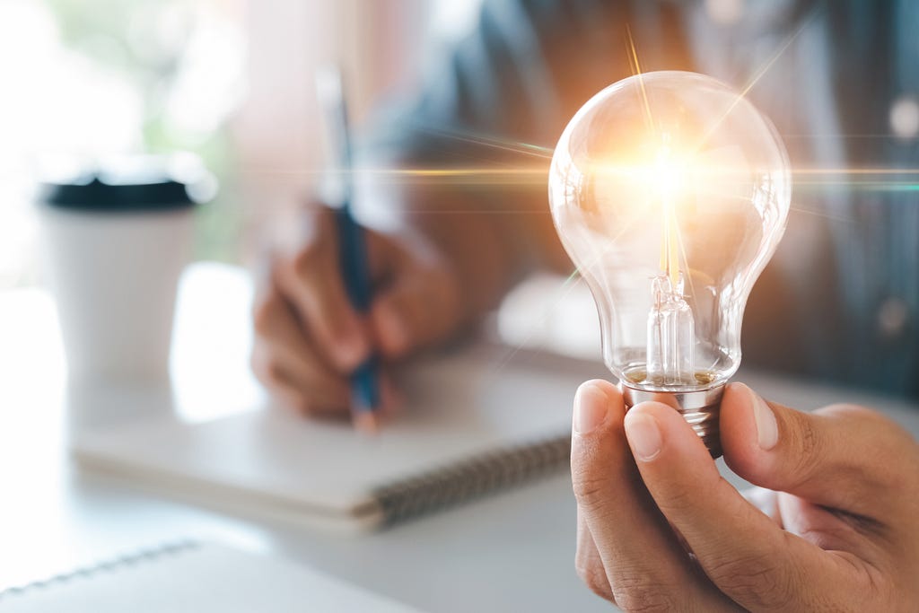 A person writing on a notepad holding a lightbulb.