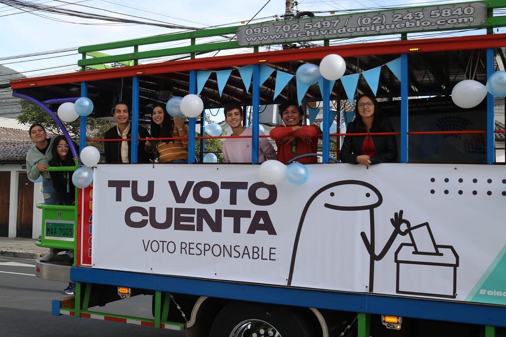 A group of people smile onboard a trolley bus that has been decorated with pennant flags and balloons, and includes a banner that reads “TU VOTO CUENTA; VOTO RESPONSABLE.”