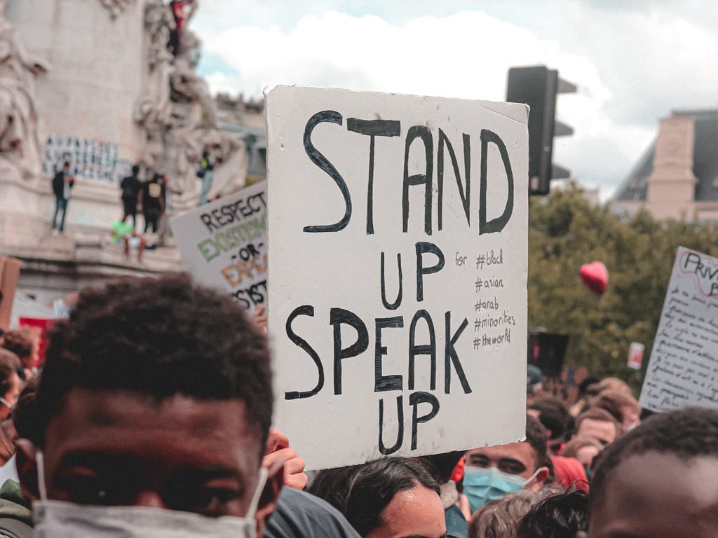 A crowd protesting with sign that says “Stand Up, Speak Up”