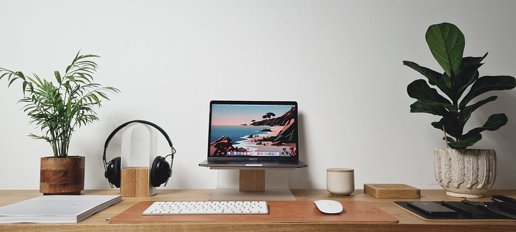 Wooden desk with a laptop perched on a stand, headphones, white wireless keyboard, and a white wireless mouse