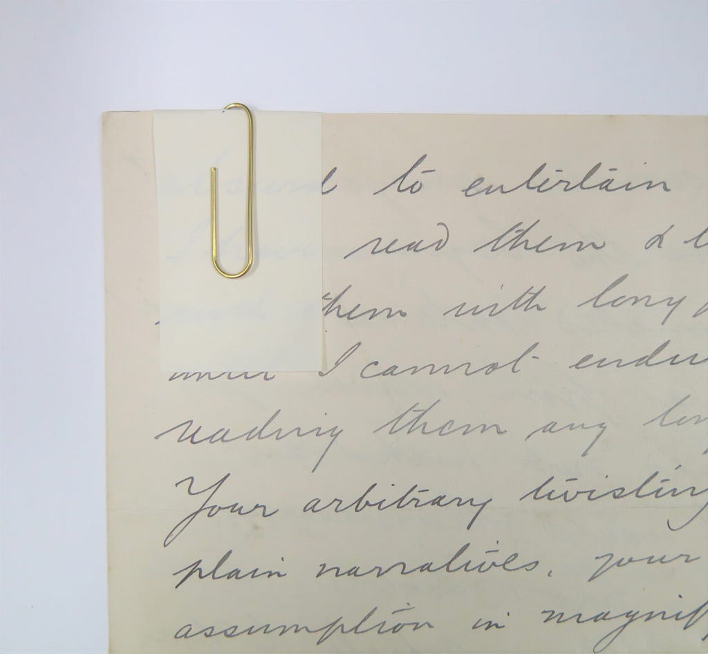 A close up of the top left hand corner of handwritten documents which are held together with an archival brass paperclip over a tab of folded archival paper.